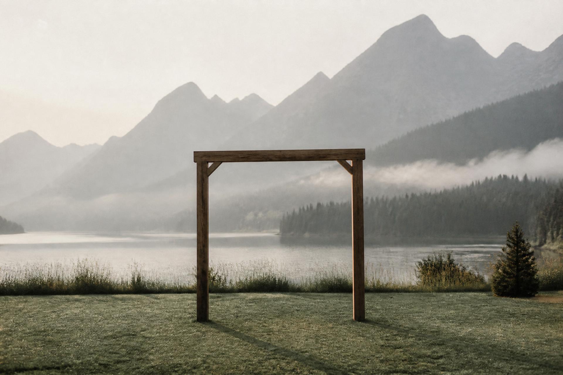 Bare wooden ceremony arch on a misty lawn beside a mountain lake at dawn